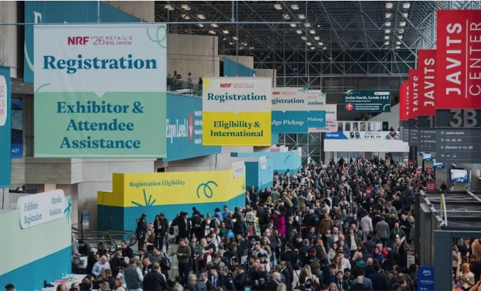 Uma visão ampla do interior do Javits Center repleto de milhares de participantes. No teto alto, pendem vários banners grandes de sinalização para o evento 'NRF '26 Retail's Big Show'. Um banner verde indica 'Registration: Exhibitor & Attendee Assistance' e um amarelo indica 'Eligibility & International'. À direita, faixas verticais vermelhas mostram o nome 'JAVITS CENTER'.