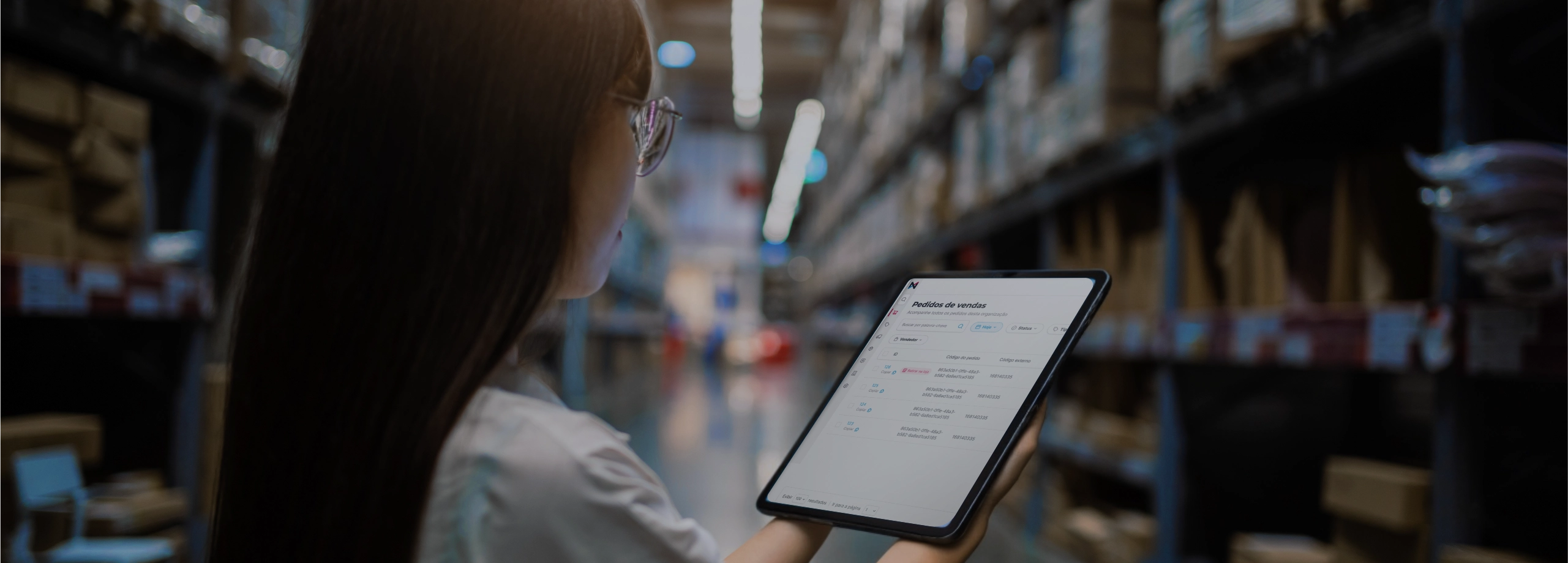 Visto por cima do ombro, uma mulher de cabelos longos e óculos segura um tablet com as duas mãos. A tela exibe uma lista de pedidos ou produtos. Ela está parada no corredor central de um depósito amplo, cercada por prateleiras industriais altas repletas de caixas de papelão em ambos os lados.