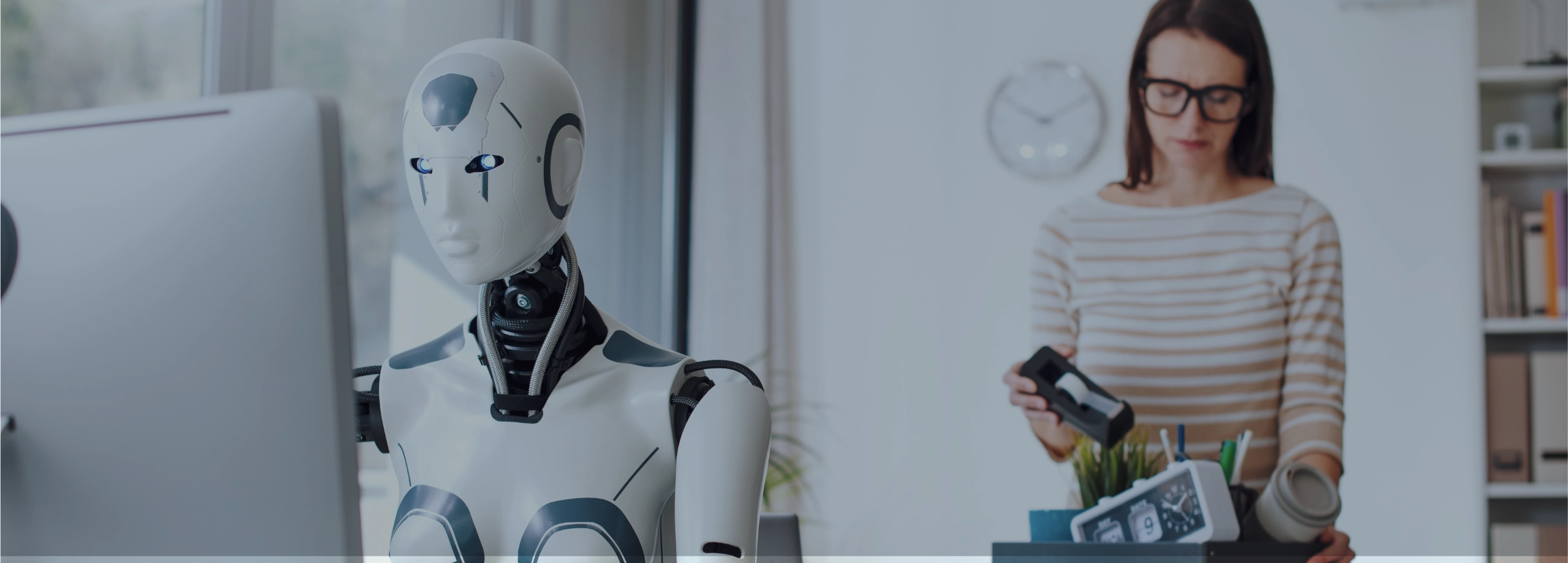 Cena em um escritório: à esquerda e em foco, um robô branco com traços humanos está sentado digitando em um computador. Ao fundo, levemente desfocada, uma mulher de óculos e blusa listrada está em pé, guardando itens de mesa (como um relógio e porta-canetas) dentro de uma caixa preta, sugerindo que ela está deixando o emprego ou sendo substituída.