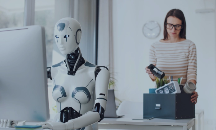 Cena em um escritório: à esquerda e em foco, um robô branco com traços humanos está sentado digitando em um computador. Ao fundo, levemente desfocada, uma mulher de óculos e blusa listrada está em pé, guardando itens de mesa (como um relógio e porta-canetas) dentro de uma caixa preta, sugerindo que ela está deixando o emprego ou sendo substituída.
