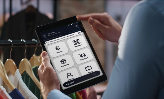 Close-up das mãos de um funcionário de camisa azul segurando um tablet. A tela exibe o painel de um aplicativo de varejo com botões grandes para: Vendas, Bipagem, Produtos, Retiradas, Clientes e Troca. No topo da tela, lê-se 'Olá, João'. O fundo está desfocado, mostrando cabides de madeira com roupas em uma arara.