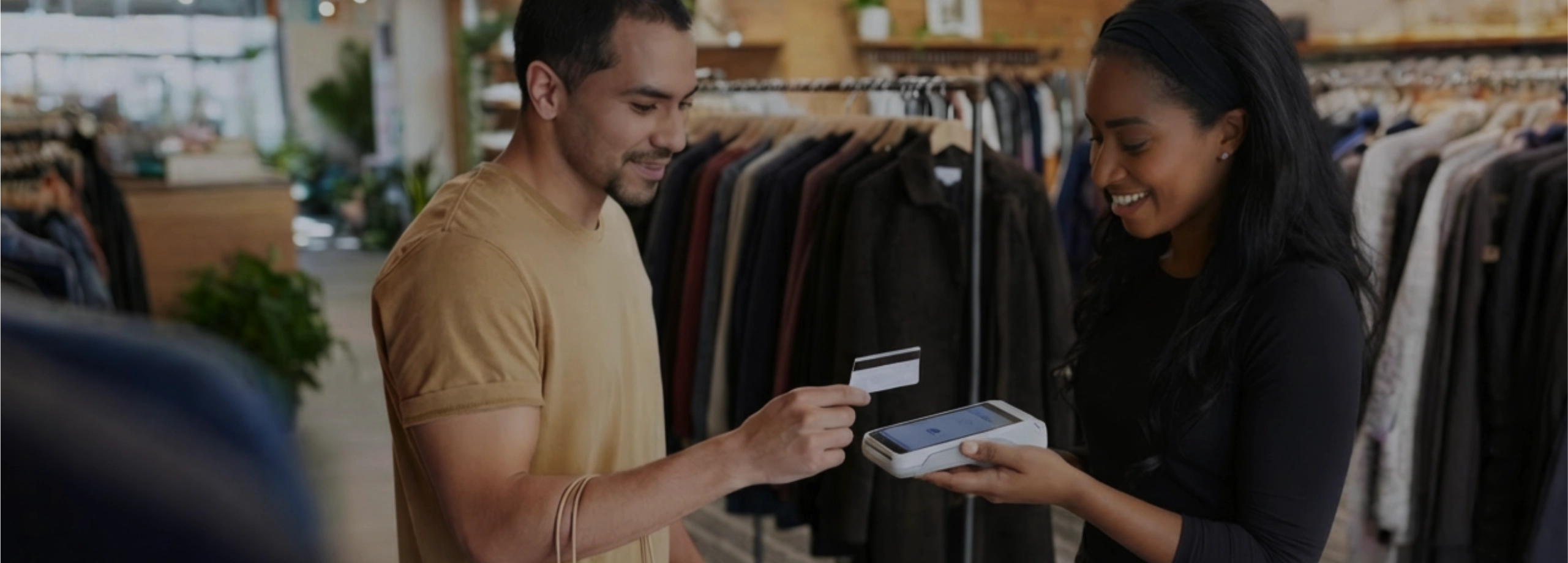 Cena em uma loja de roupas: à esquerda, um cliente de camiseta mostarda segura uma sacola de papel e estende um cartão de crédito. À direita, uma vendedora negra, vestida de preto e usando uma faixa no cabelo, sorri enquanto segura um terminal de pagamento branco (Smart POS) para processar a venda. O fundo mostra o interior da loja com roupas organizadas em araras.