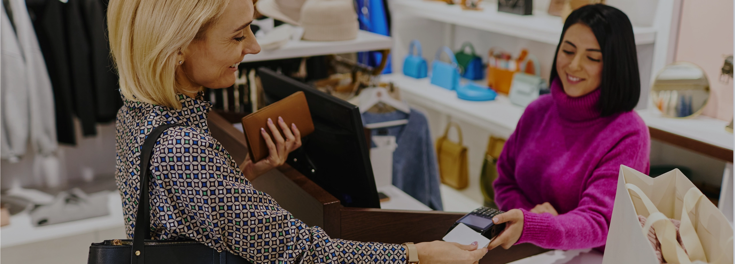 Cena no caixa de uma loja de moda. À esquerda, uma cliente loira de blusa estampada e bolsa preta no ombro sorri ao inserir um cartão branco no terminal de pagamento. Na outra mão, ela segura uma carteira marrom. À direita, a vendedora, vestindo um suéter rosa vibrante, segura a maquininha e sorri de volta. No balcão branco entre elas, vê-se as costas do monitor do caixa e uma sacola de papel. Ao fundo, prateleiras iluminadas exibem bolsas coloridas e araras com roupas.