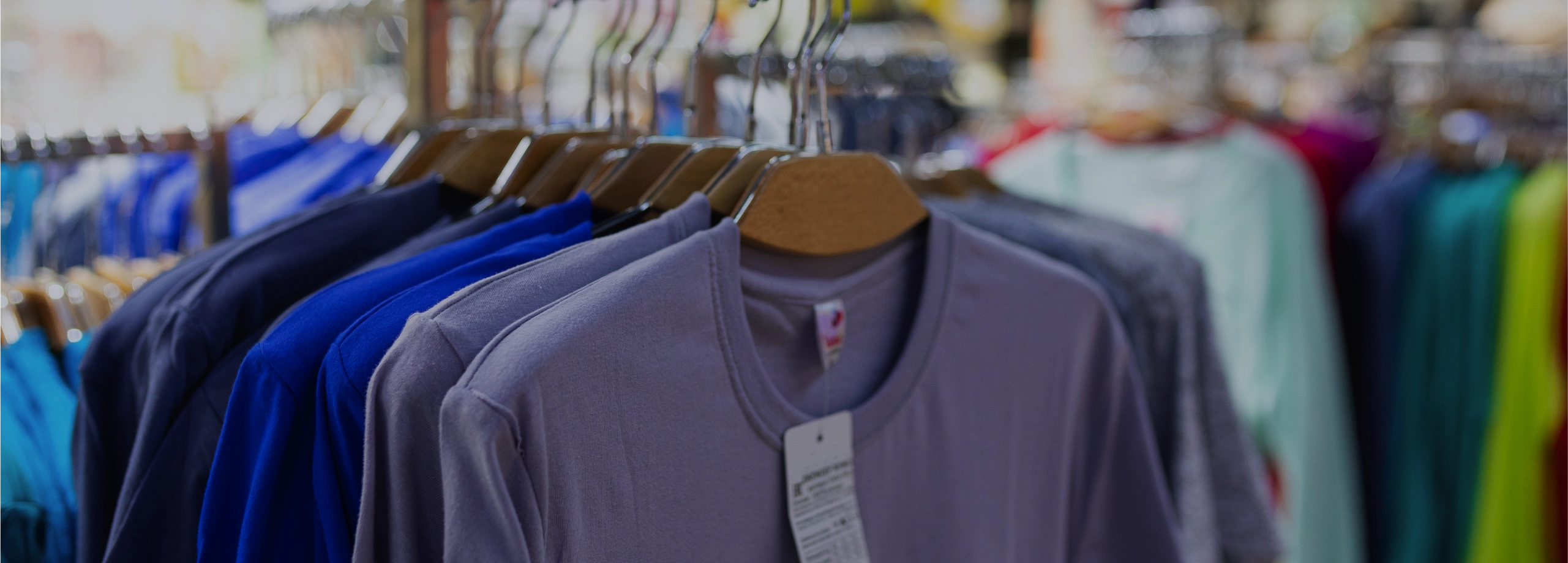 Close-up de uma camiseta cinza com etiqueta pendurada em um cabide de madeira, em uma arara com outras roupas coloridas dentro de uma loja.