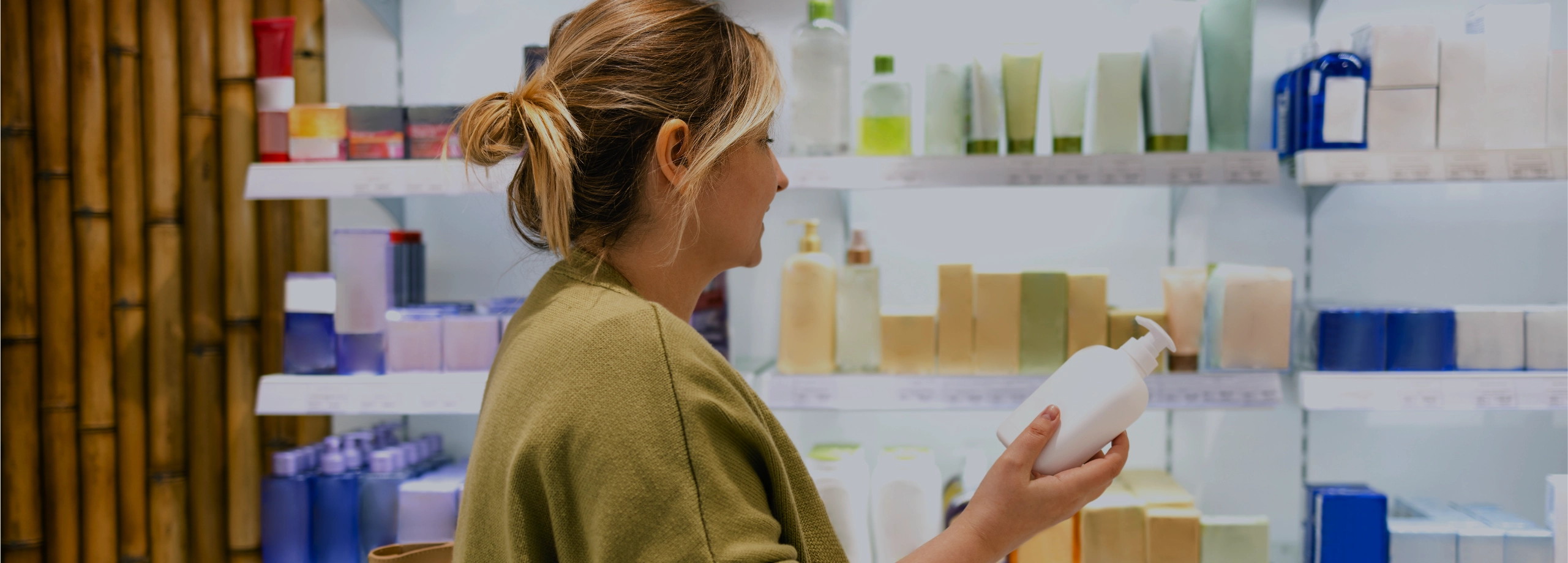 Mulher de perfil examinando um frasco branco de cosmético em frente a prateleiras repletas de produtos de beleza em uma loja.