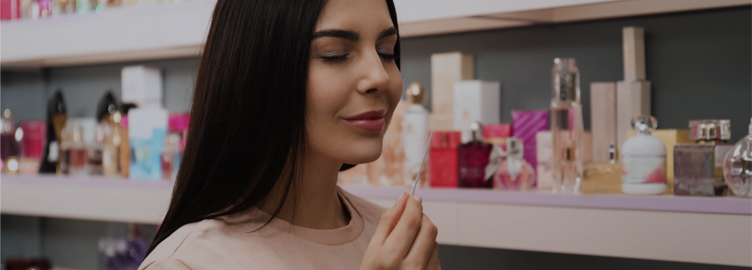 Mulher de cabelos escuros e olhos fechados sorri enquanto sente o aroma de uma fita de teste em uma perfumaria.