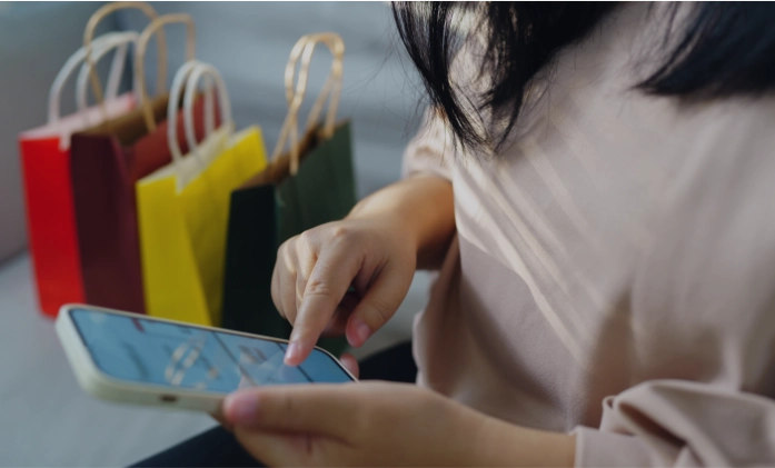 Close-up de uma pessoa usando um smartphone com várias sacolas de compras coloridas ao fundo.
