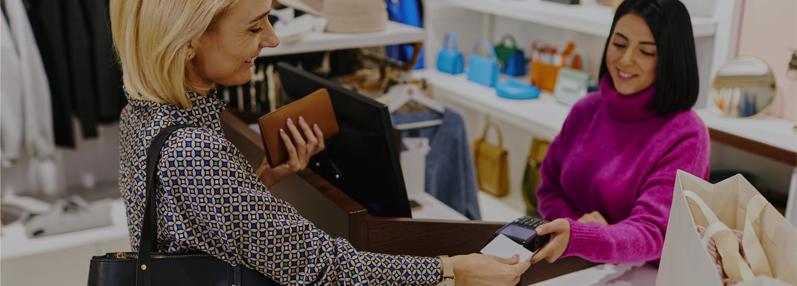 Uma cliente loira pagando com cartão em uma loja de roupas, sendo atendida por uma vendedora de blusa roxa.