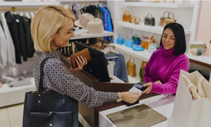Uma cliente loira pagando com cartão em uma loja de roupas, sendo atendida por uma vendedora de blusa roxa.