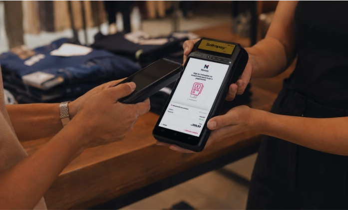 Cliente realiza pagamento por aproximação usando um smartphone em uma maquininha de cartão inteligente segurada por um vendedor.