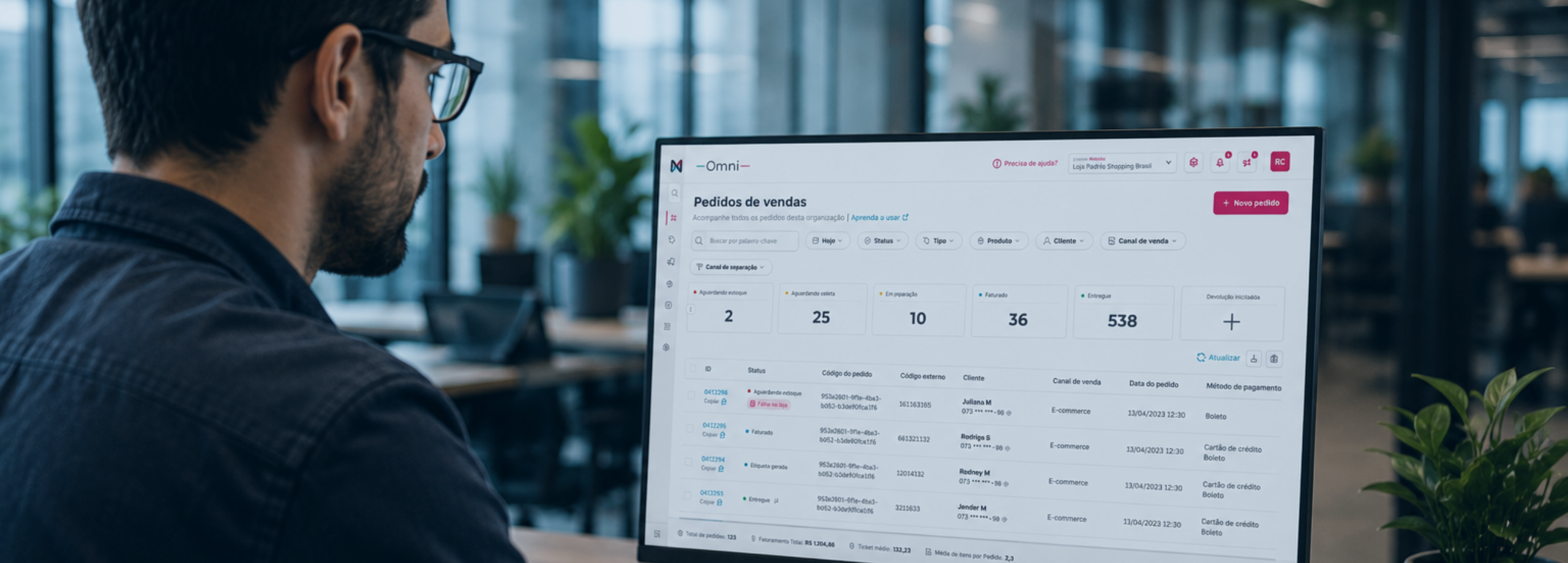 Um homem de óculos trabalhando em um escritório enquanto visualiza um painel de gerenciamento de pedidos de vendas em um monitor de computador.