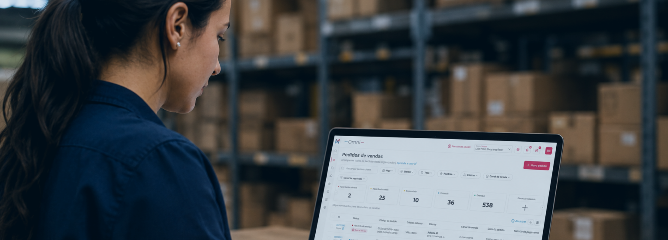 Mulher usando um notebook com um sistema de gestão de pedidos aberto, em um centro de distribuição com prateleiras cheias de caixas ao fundo.
