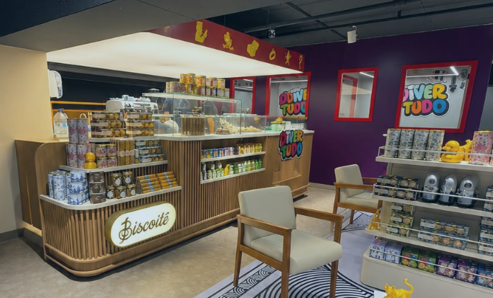 Fotografia do interior de uma loja conceito exibindo o balcão iluminado do café Biscoitê em madeira, poltronas claras e prateleiras com brinquedos e produtos sob a marca "Divertudo".