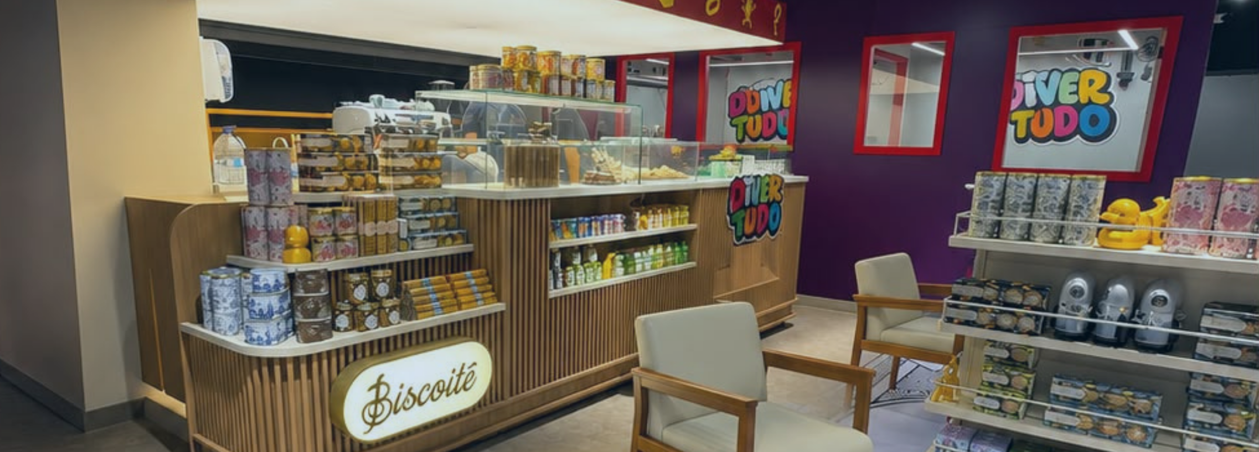 Fotografia do interior de uma loja conceito exibindo o balcão iluminado do café Biscoitê em madeira, poltronas claras e prateleiras com brinquedos e produtos sob a marca "Divertudo".