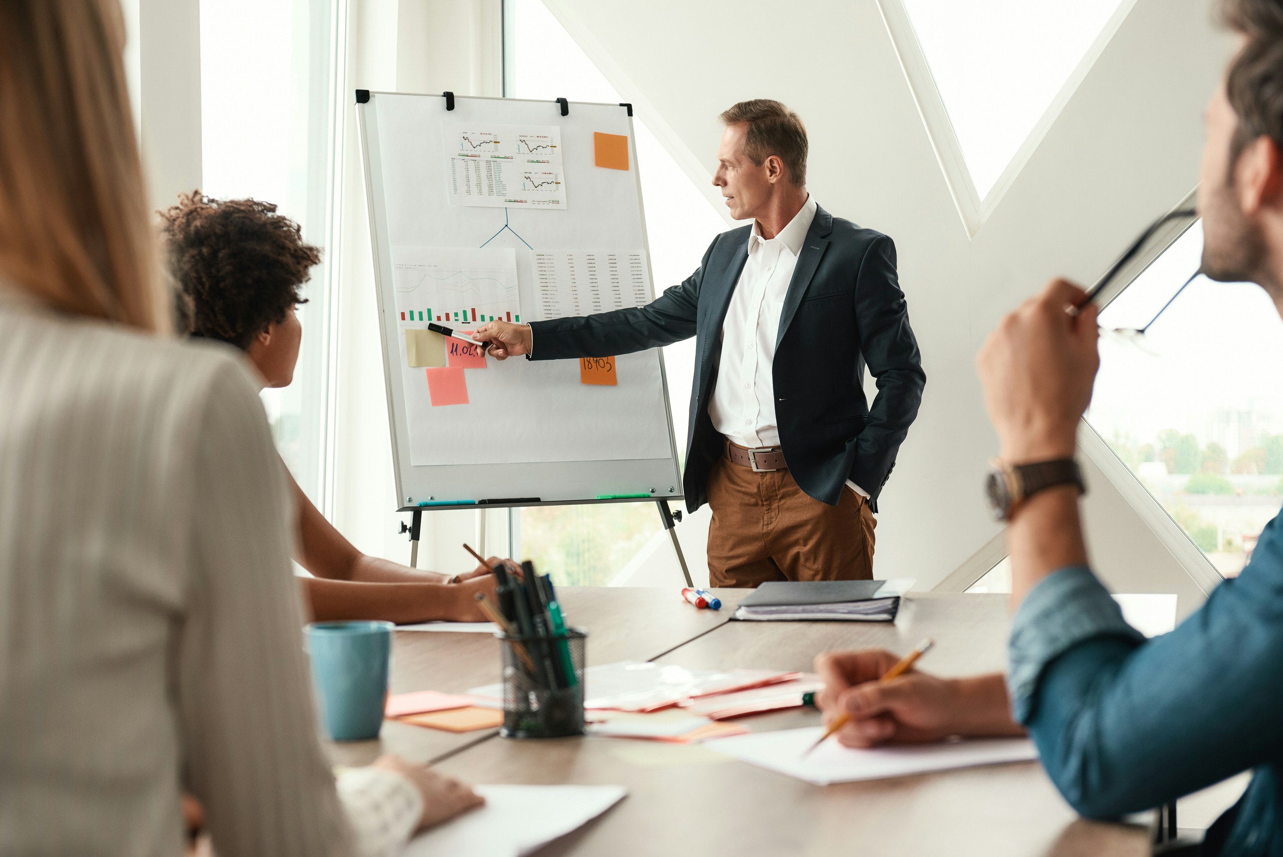 Businessman in a blazer presenting charts on a flip chart to three colleagues seated at a table in a modern office.