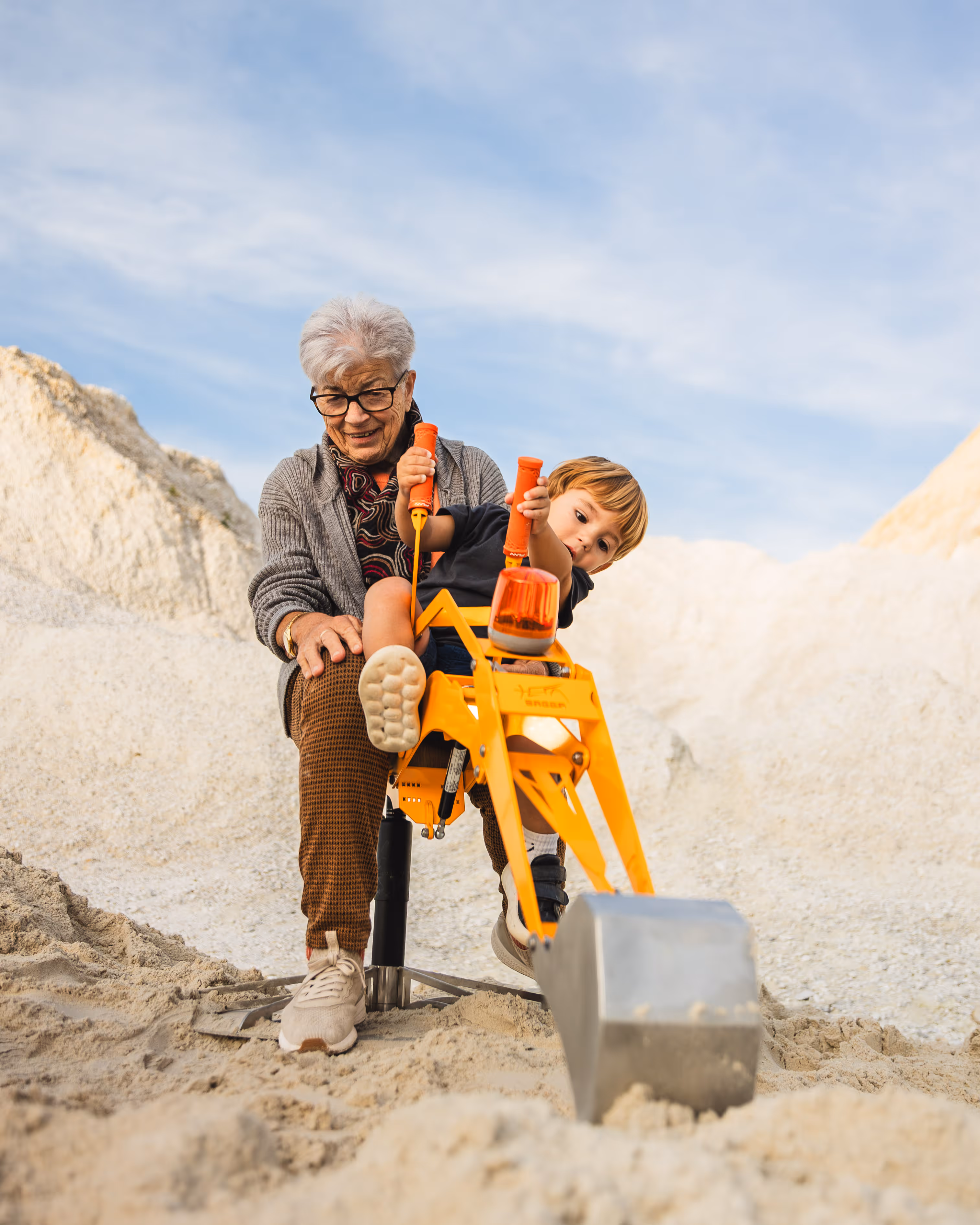 Ältere Frau sitzt mit kleinem Jungen auf einem Spielbagger und sie graben im Sand.