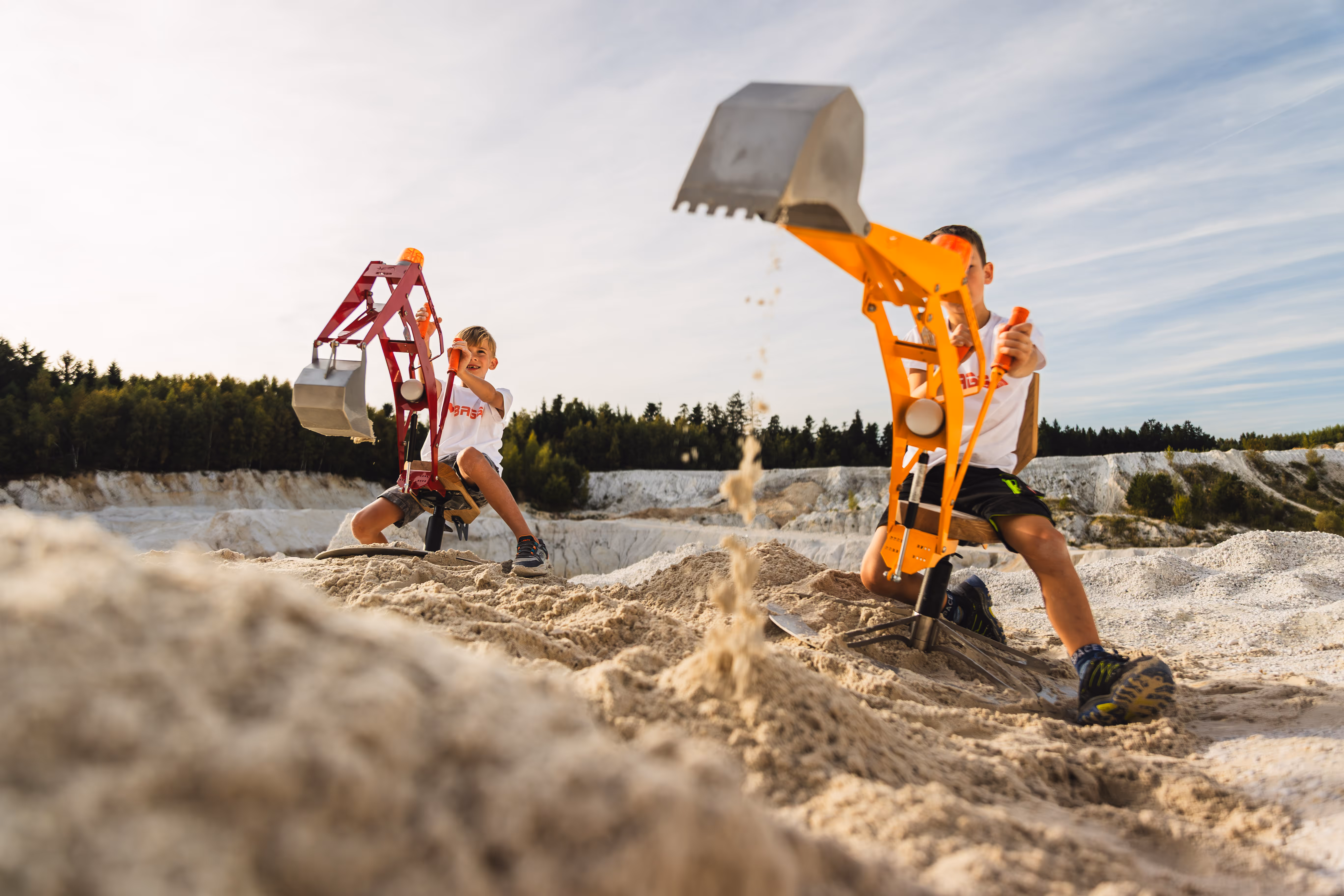 Zwei Kinder spielen mit roten und gelben Baggern in einer Sandgrube bei sonnigem Himmel.