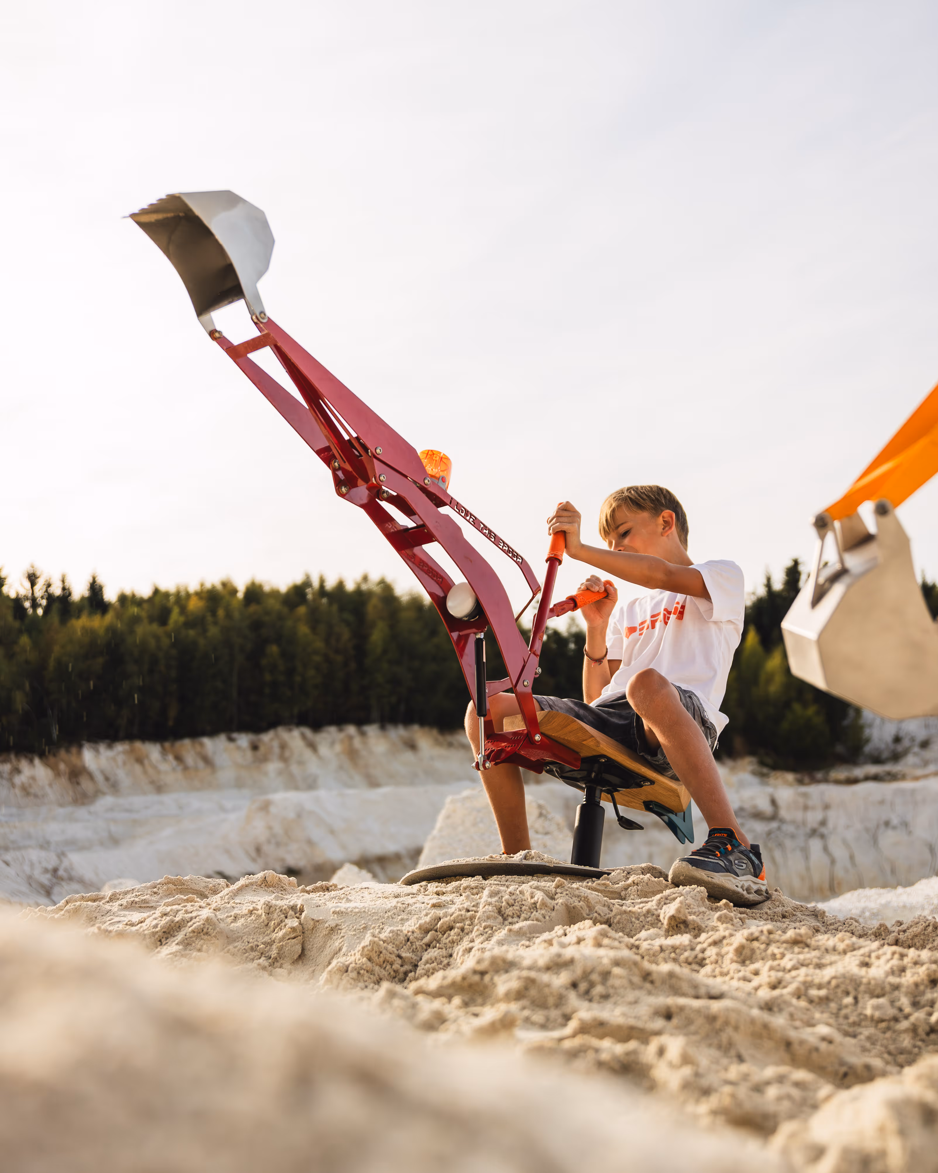 Junge sitzt auf einem Spielbagger und bewegt den roten Schaufelarm in der Luft.