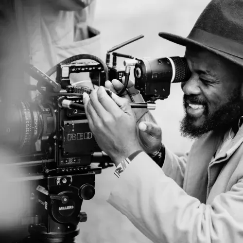 A black and white photo of a man looking into a cinema camera on a feature film set. He is smiling as he frames his shot through the viewfinder.