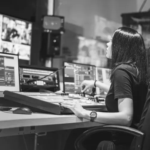 A black and white photo of a woman working on post-production for a specialty project. She is sitting at a desk using a computer, facing several monitors.