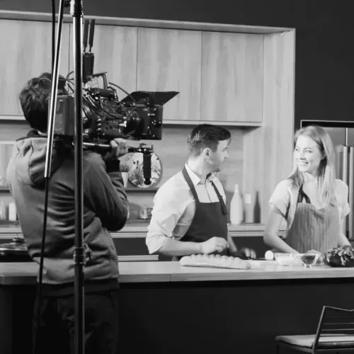 A black and white photo of an episodic TV set showing two cast members in a kitchen. A camera operator in the foreground is filming them.