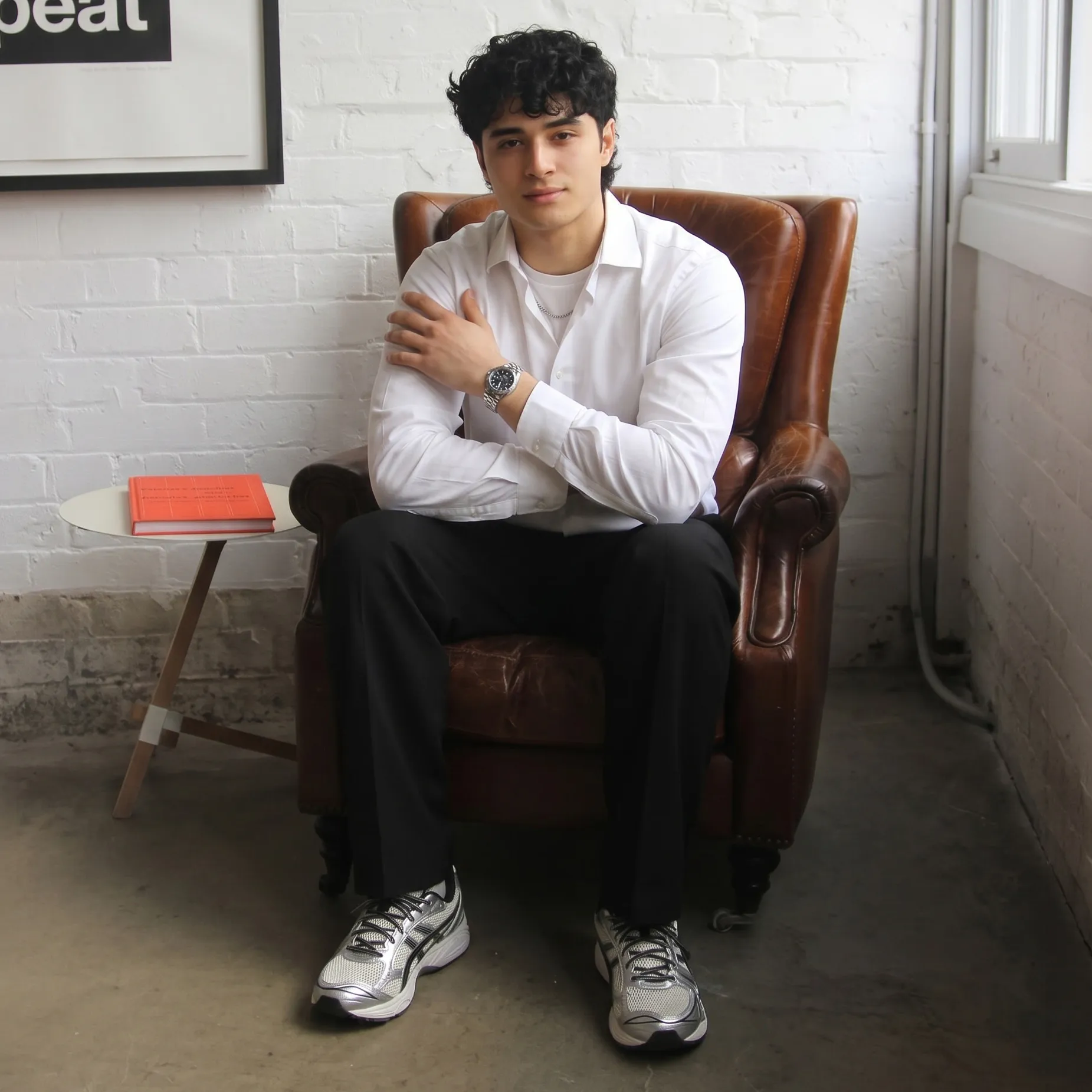 Young man in white shirt and black pants sitting on a brown leather chair by a white brick wall.