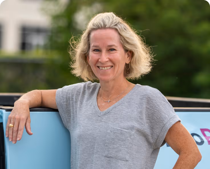 Smiling blonde woman with short hair wearing a gray shirt, leaning on a light blue surface with greenery in the background.