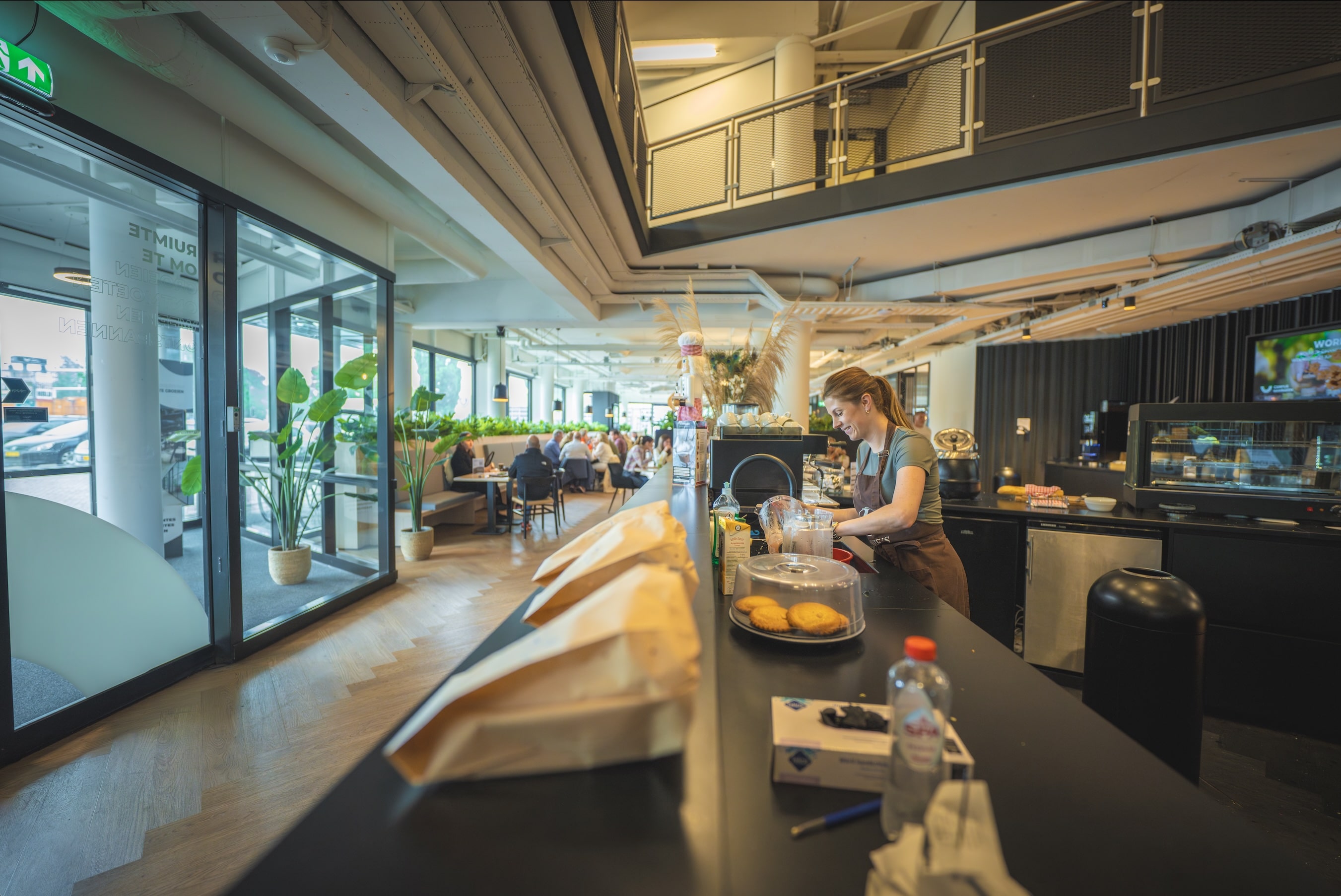 Moderne en sfeervolle horecaruimte in een bedrijfsomgeving met een open balie, bloemdecoratie en mensen die samen lunchen of overleggen aan ronde tafels in een lichte, uitnodigende setting.