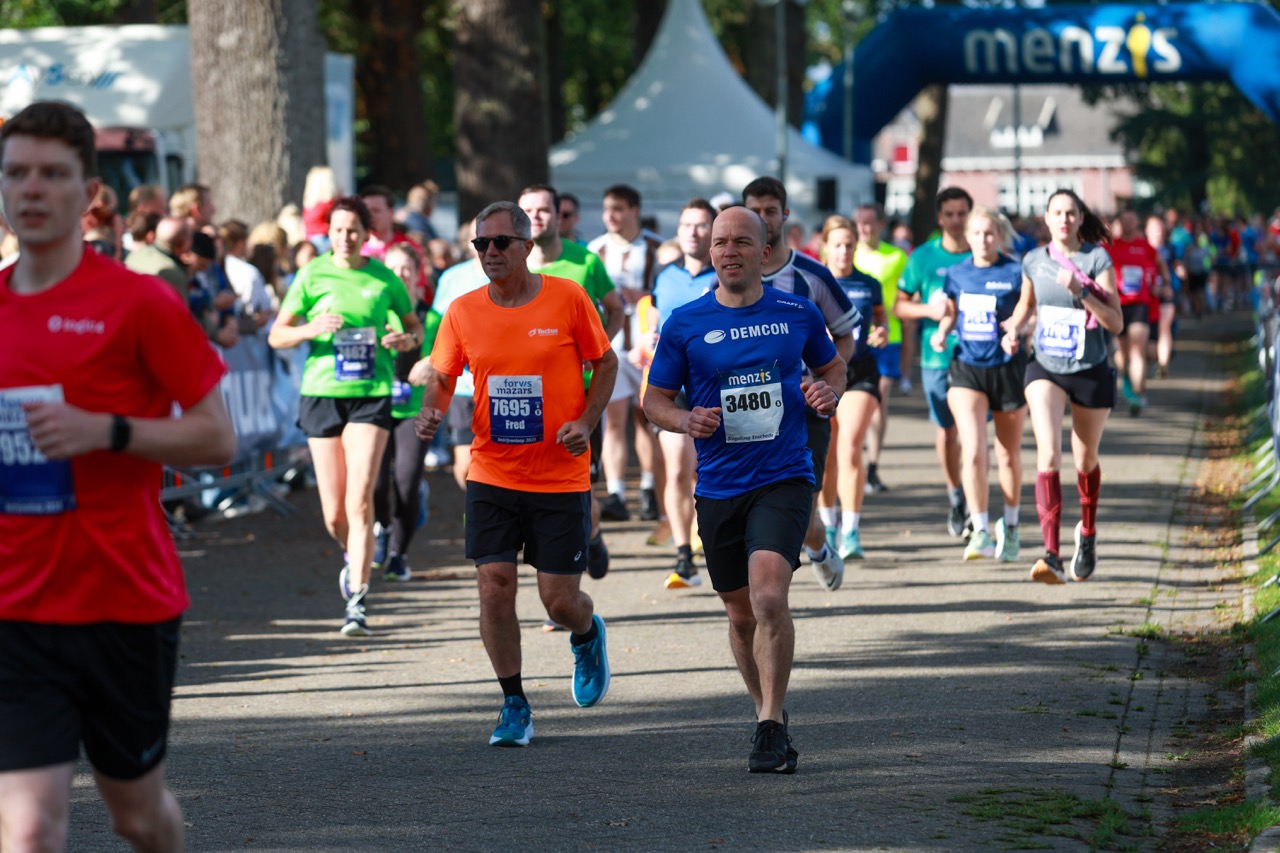 Grote groep hardlopers tijdens de Singelloop Enschede, met meerdere deelnemers in bedrijfsshirts vooraan in beeld.