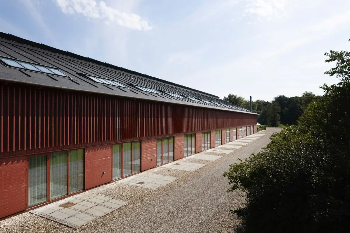 Langt rødt bindingsværkshus med store vinduer langs en grusvej med træer til højre under en blå himmel.
