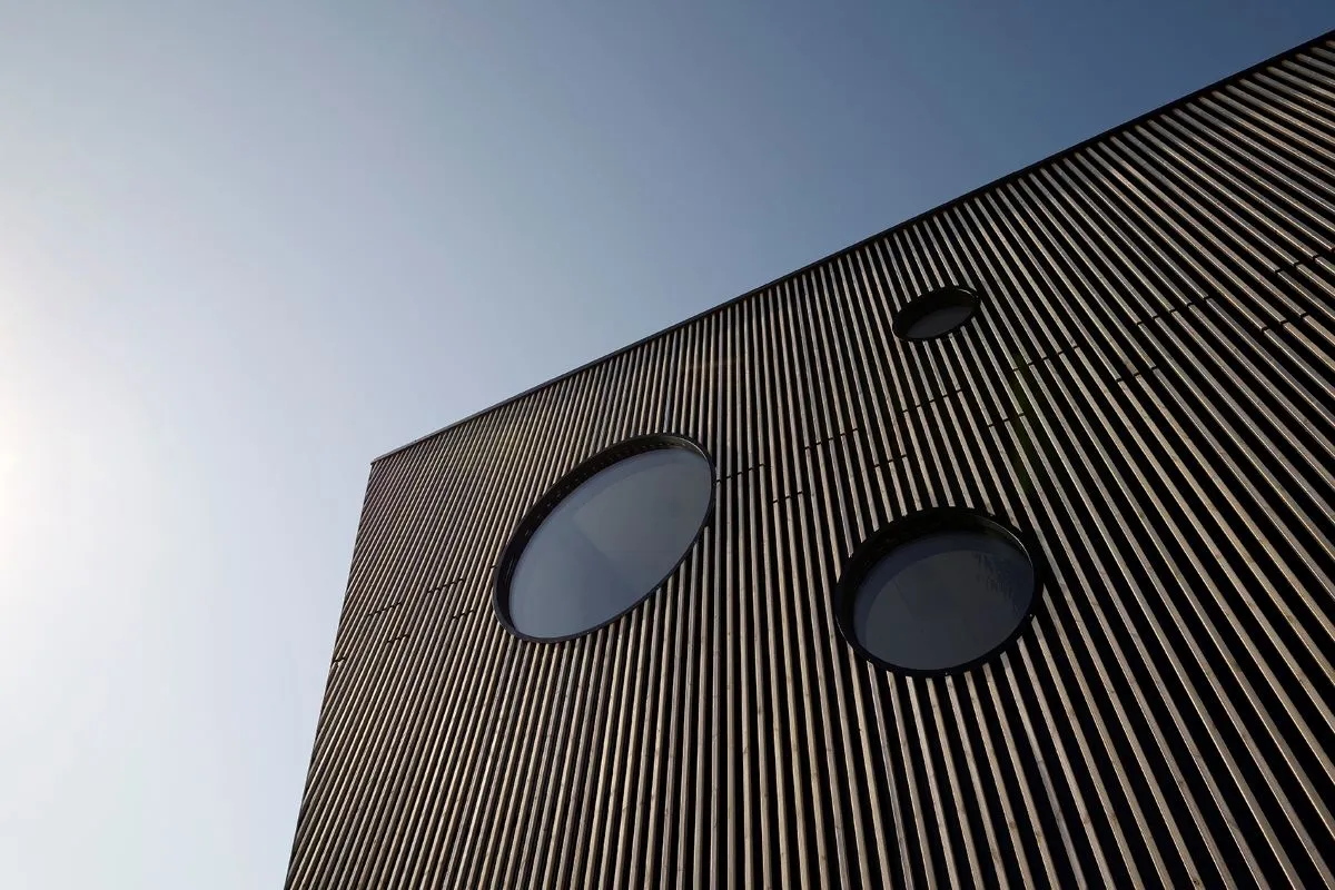 Moderne bygning med lodrette træpaneler og tre runde vinduer under en klar himmel.