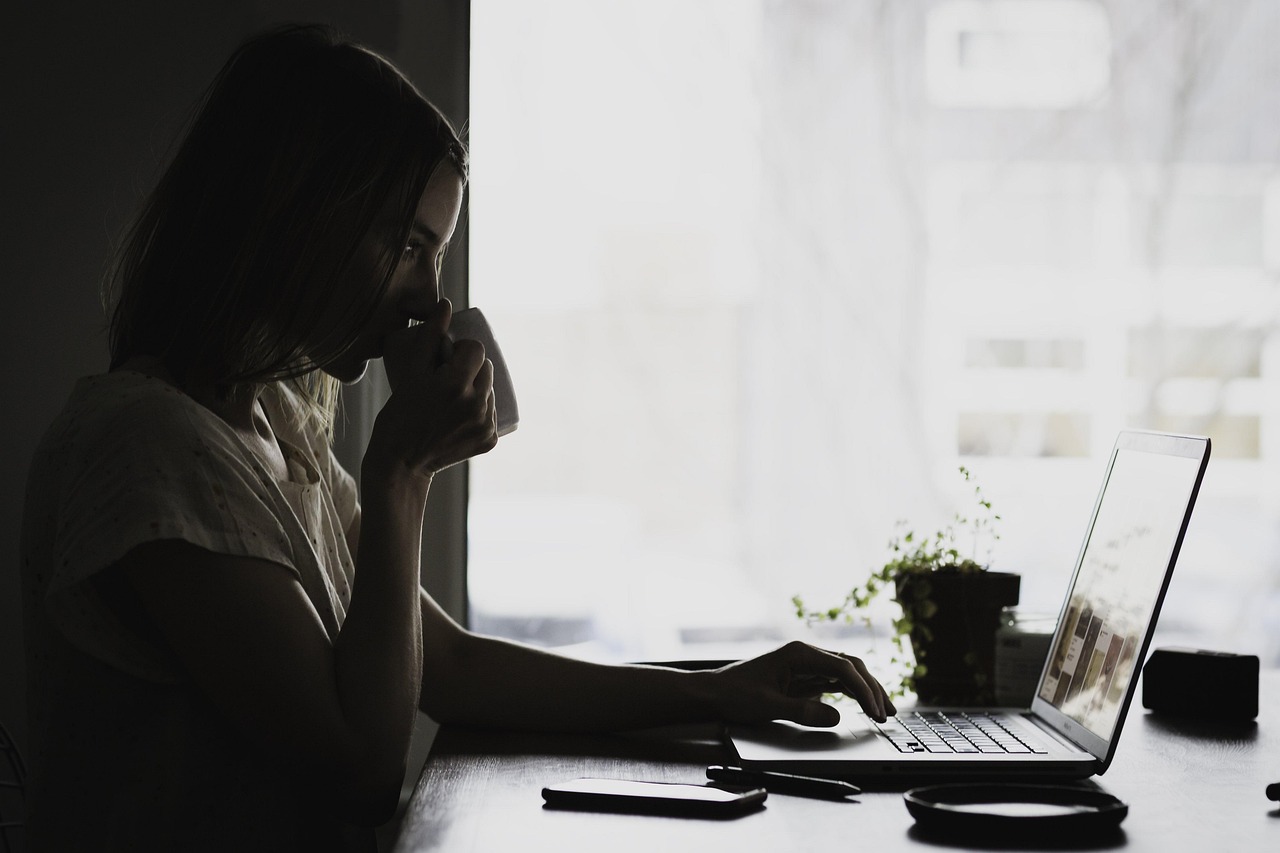 Woman working from home, drinking from a mug while using a laptop at a dimly lit desk.