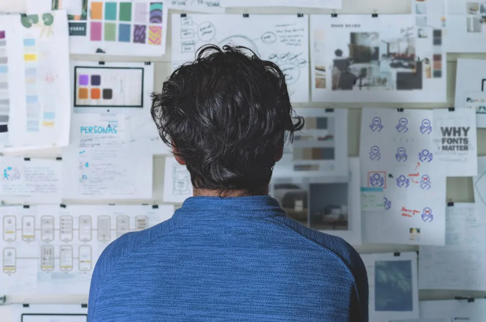A designer in a blue shirt reviewing a wall covered with creative sketches, diagrams, and notes, symbolizing brainstorming and project planning in design or development.