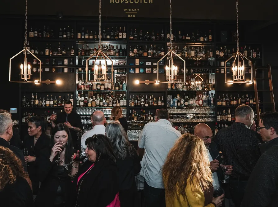 People gathered at a bar with shelves full of various bottles of alcohol, socializing under modern hanging light fixtures.