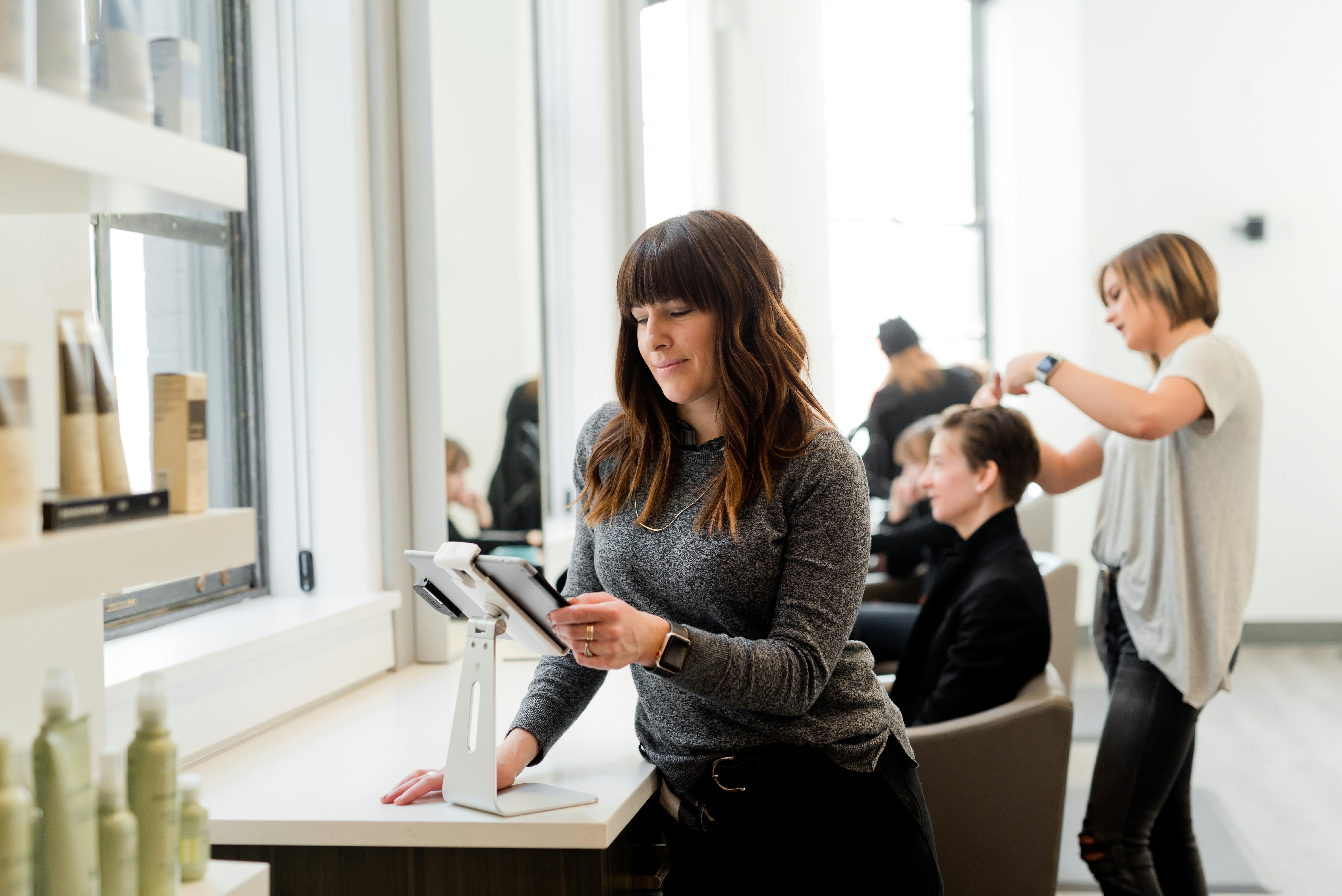 Receptionist using a tablet at a modern salon front desk in Saint John, NB, while hairstylists assist clients in the background.