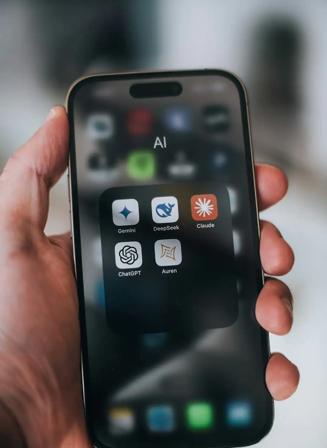 Close-up of a smartphone screen showing a folder labeled "AI" with popular AI apps including Gemini, DeepSeek, Claude, ChatGPT, and Auren.