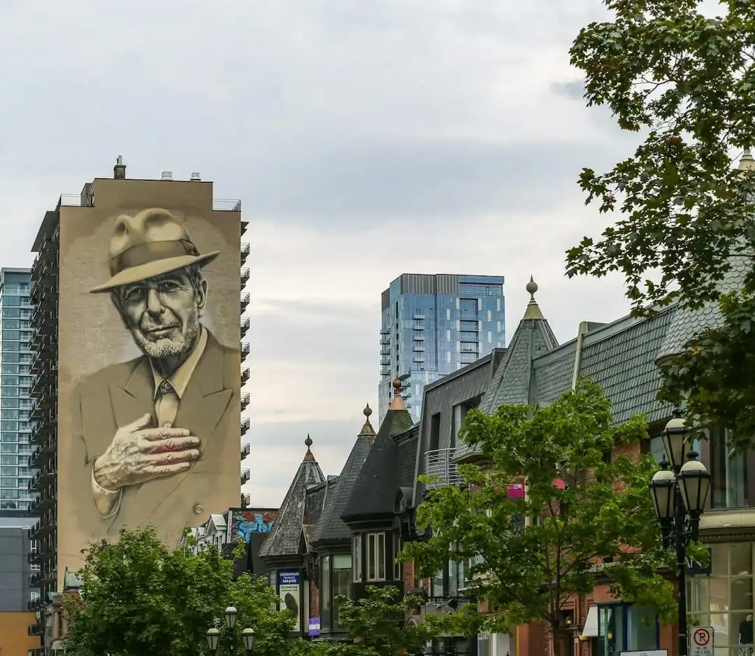 Famous mural of Leonard Cohen on the side of a building in downtown Montreal, surrounded by trees and classic architecture.