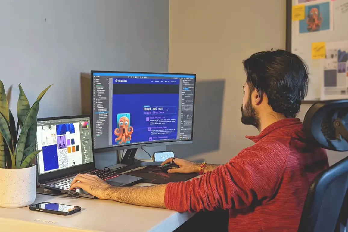 A designer working at a dual-monitor setup, with one screen showing a web design for DigiBenders on Web Flow, featuring a playful octopus illustration and the text 'Check out our case studies.' The other monitor displays design assets in a graphic design software. A potted plant sits beside the setup, and the person is focused on the work in progress.