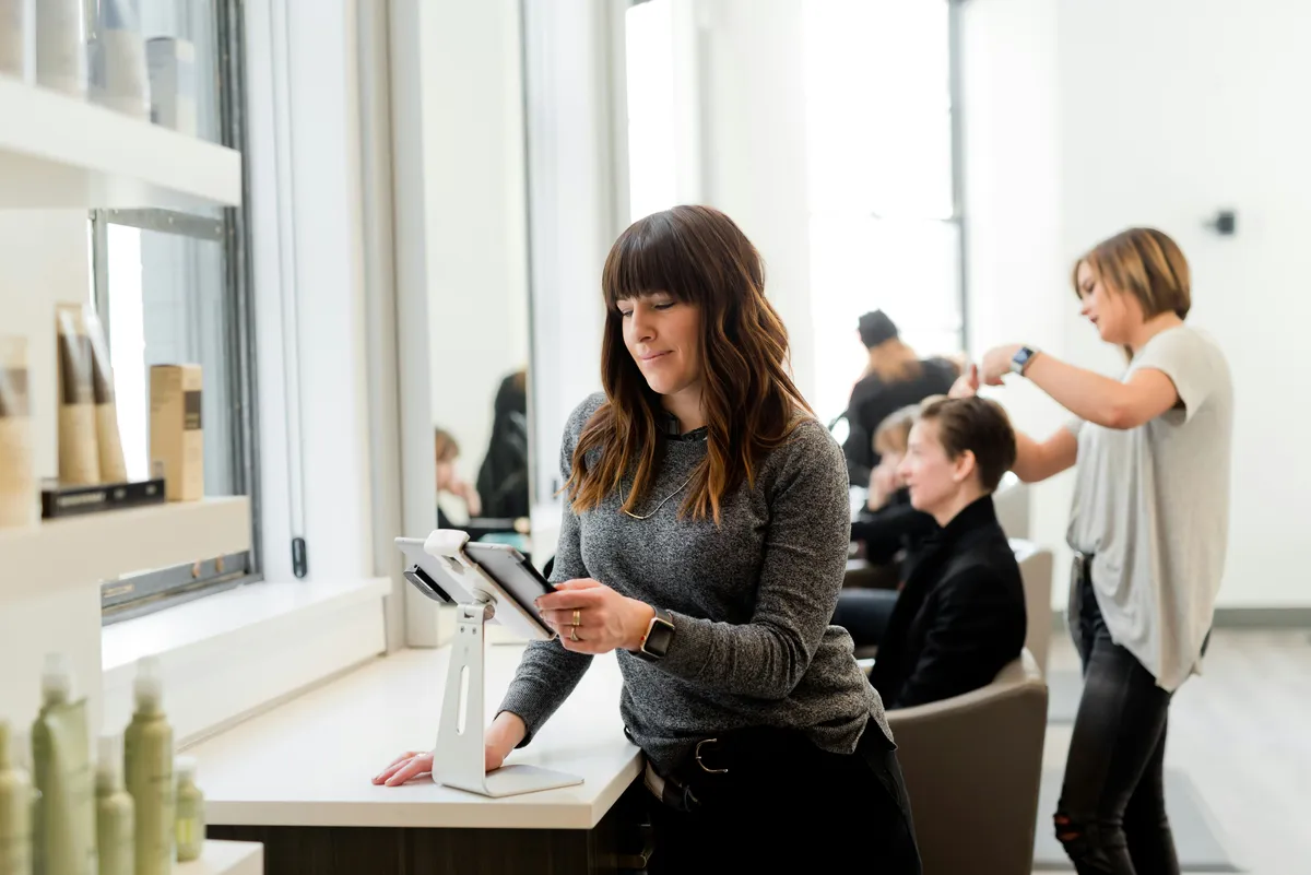 Receptionist using a tablet at a modern salon front desk in Saint John, NB, while hairstylists assist clients in the background.