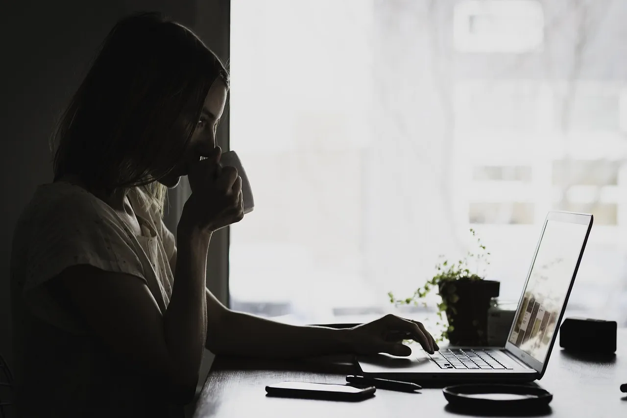 Woman working from home, drinking from a mug while using a laptop at a dimly lit desk.
