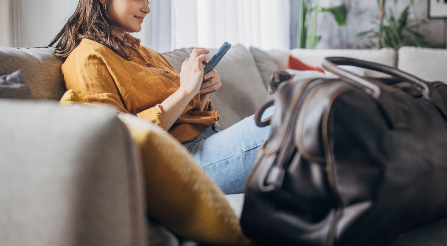 Image of a woman sitting on the couch, using her phone with her duffel by her side