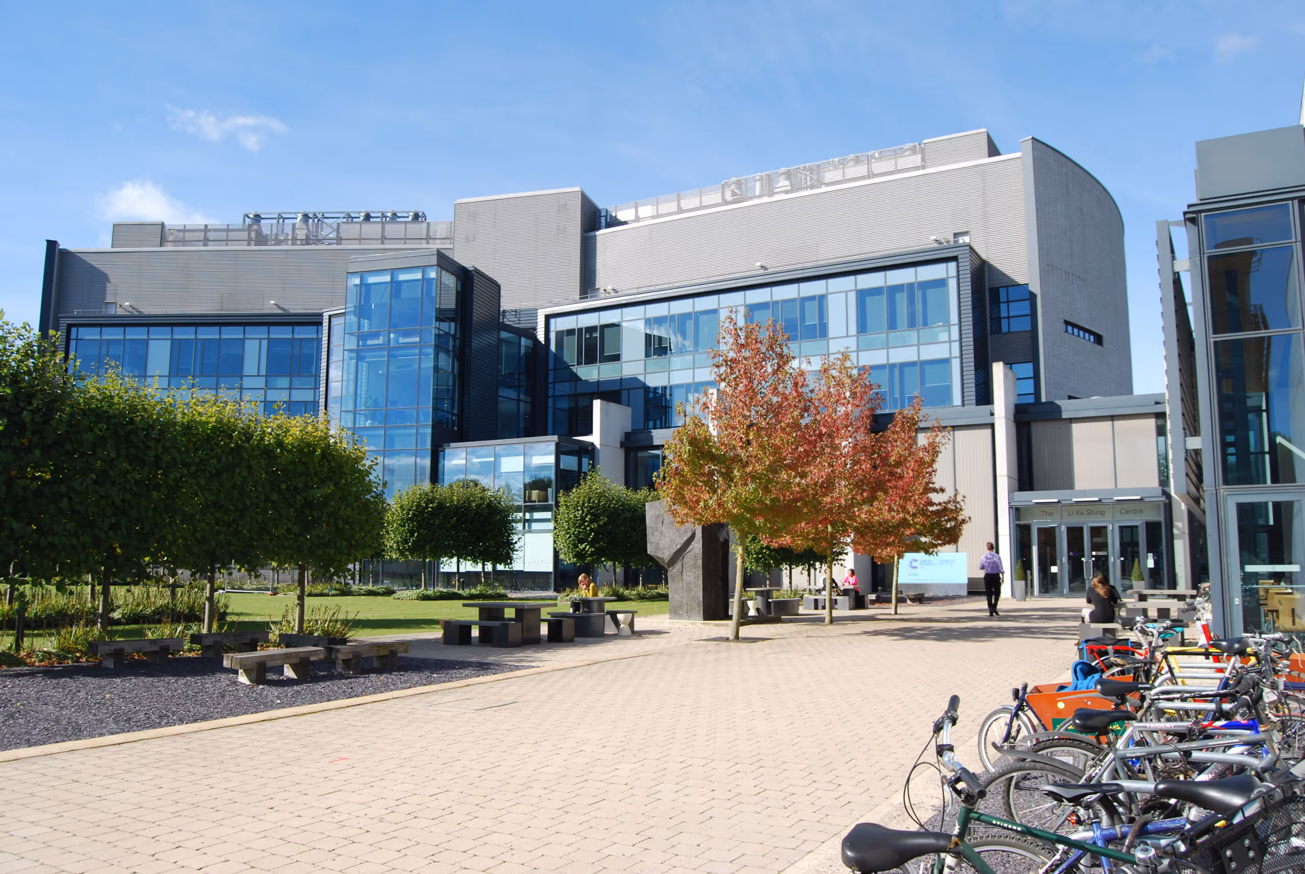 Modern building with large glass windows, surrounded by trees, benches, people, and a row of parked bicycles on a sunny day.