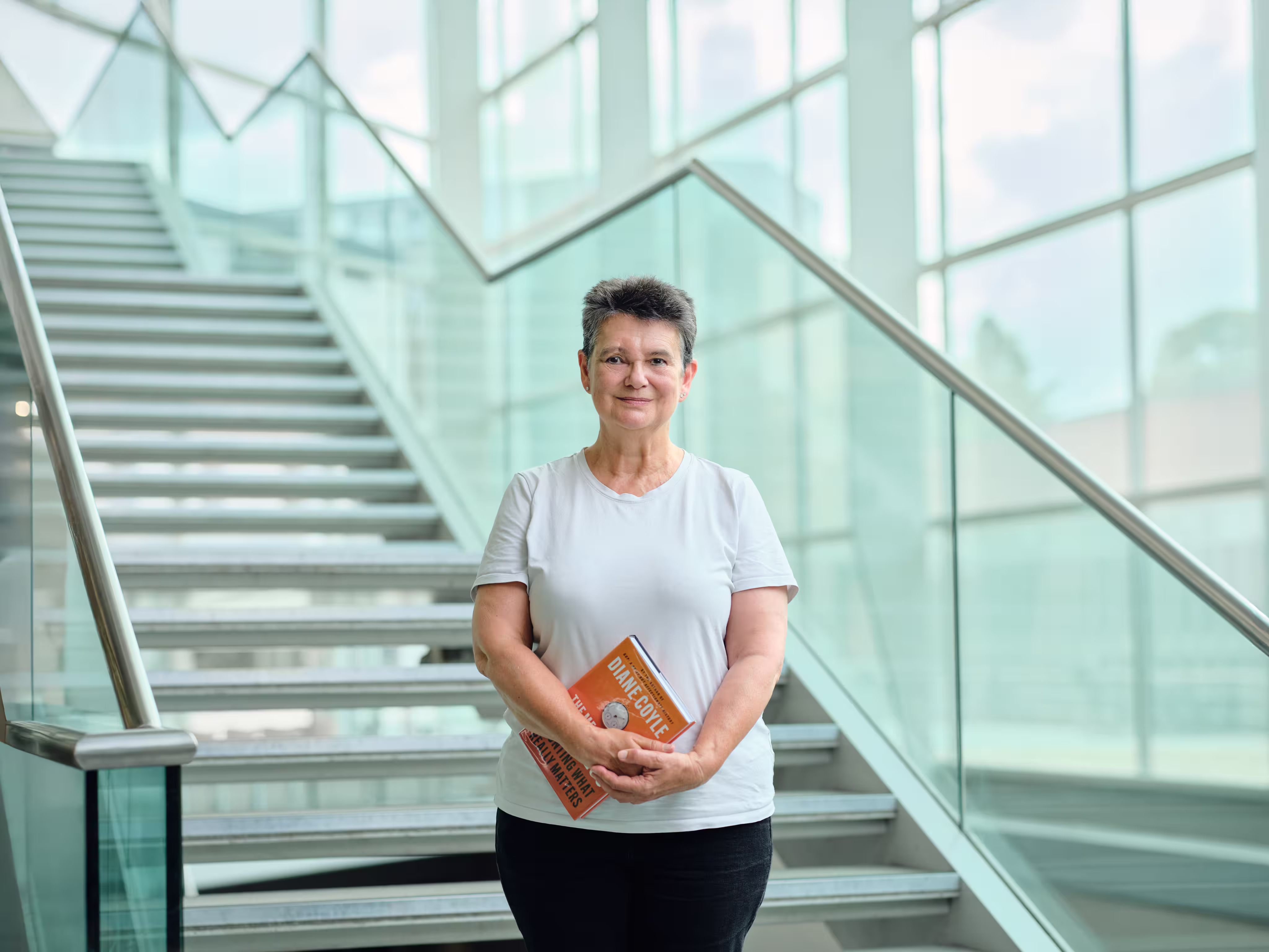 Smiling middle-aged woman in a white shirt holding a book titled ‘Diane Coyle: The Wealth of Nations’ standing in front of a glass staircase.