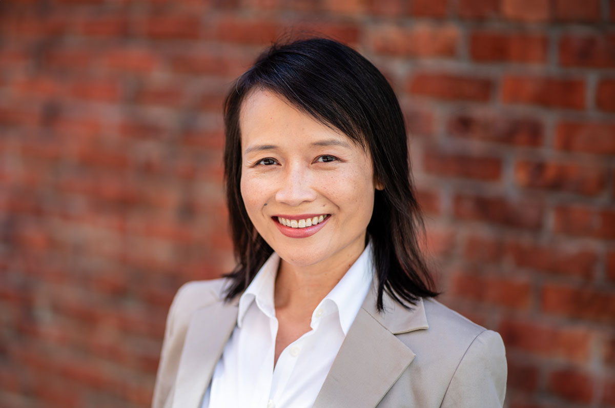 Smiling woman with short black hair wearing a white shirt and beige blazer against a blurred red brick wall.