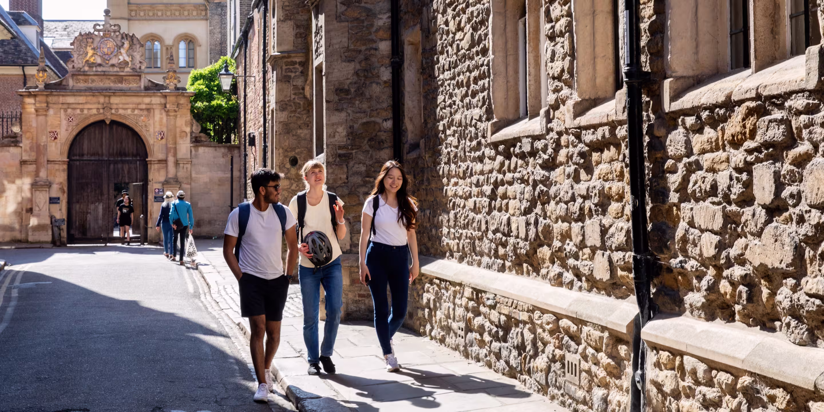 Three young people walking and talking on a sunny sidewalk next to an old stone building with arched wooden doorway in the background.