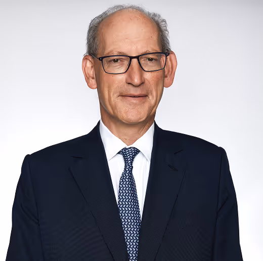 Portrait of a middle-aged man wearing glasses, a dark suit, white shirt, and patterned tie against a light background.