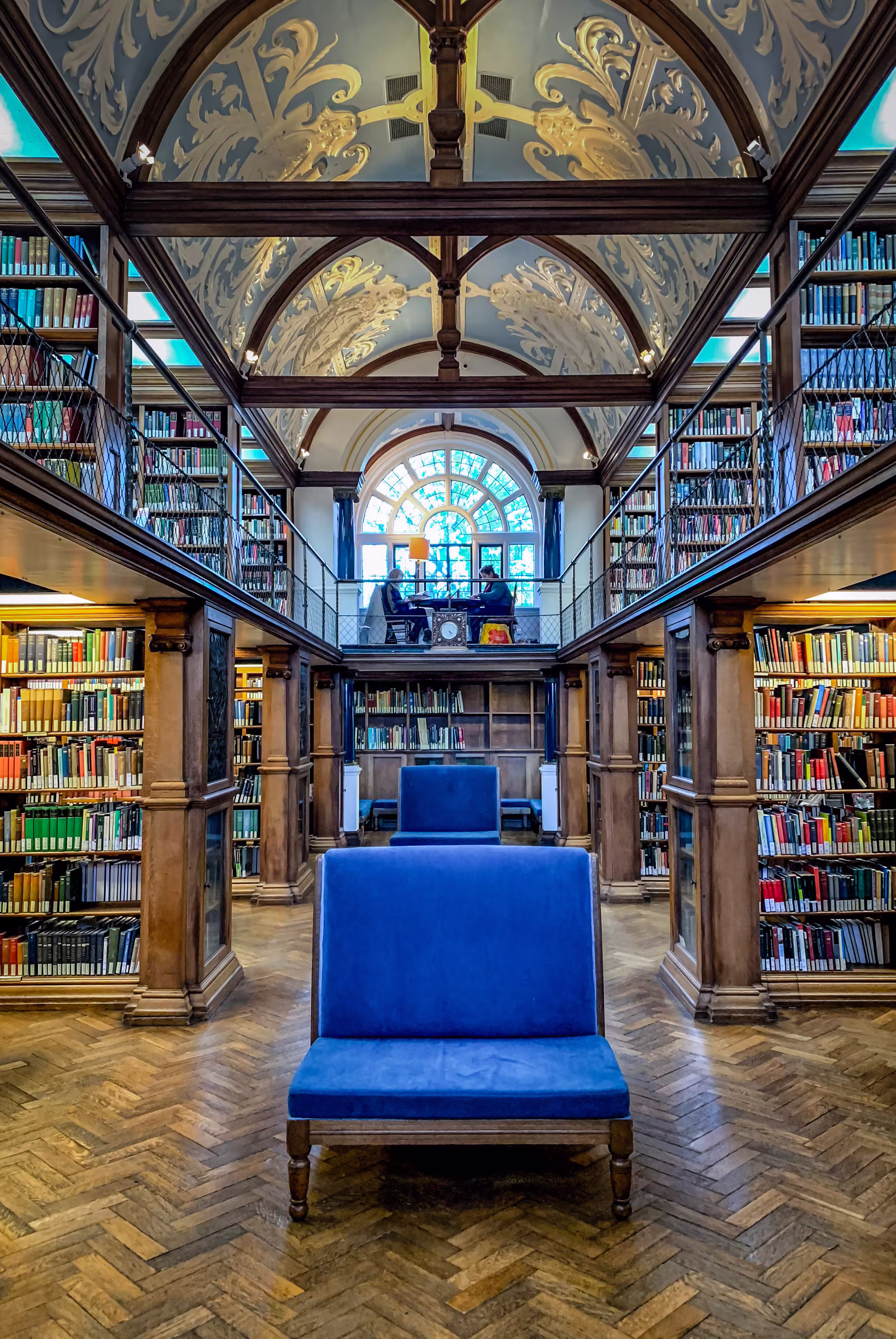 Interior of a grand library with wooden shelves filled with books, ornate arched ceiling, blue velvet benches, and two people studying by a large window.