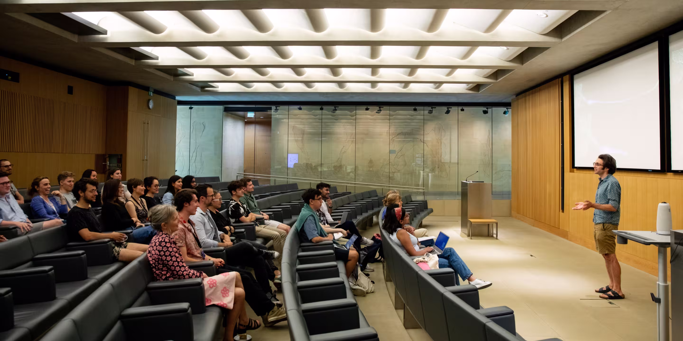 Man standing and giving a presentation to a seated audience in a modern lecture hall with wood paneling and large screens.