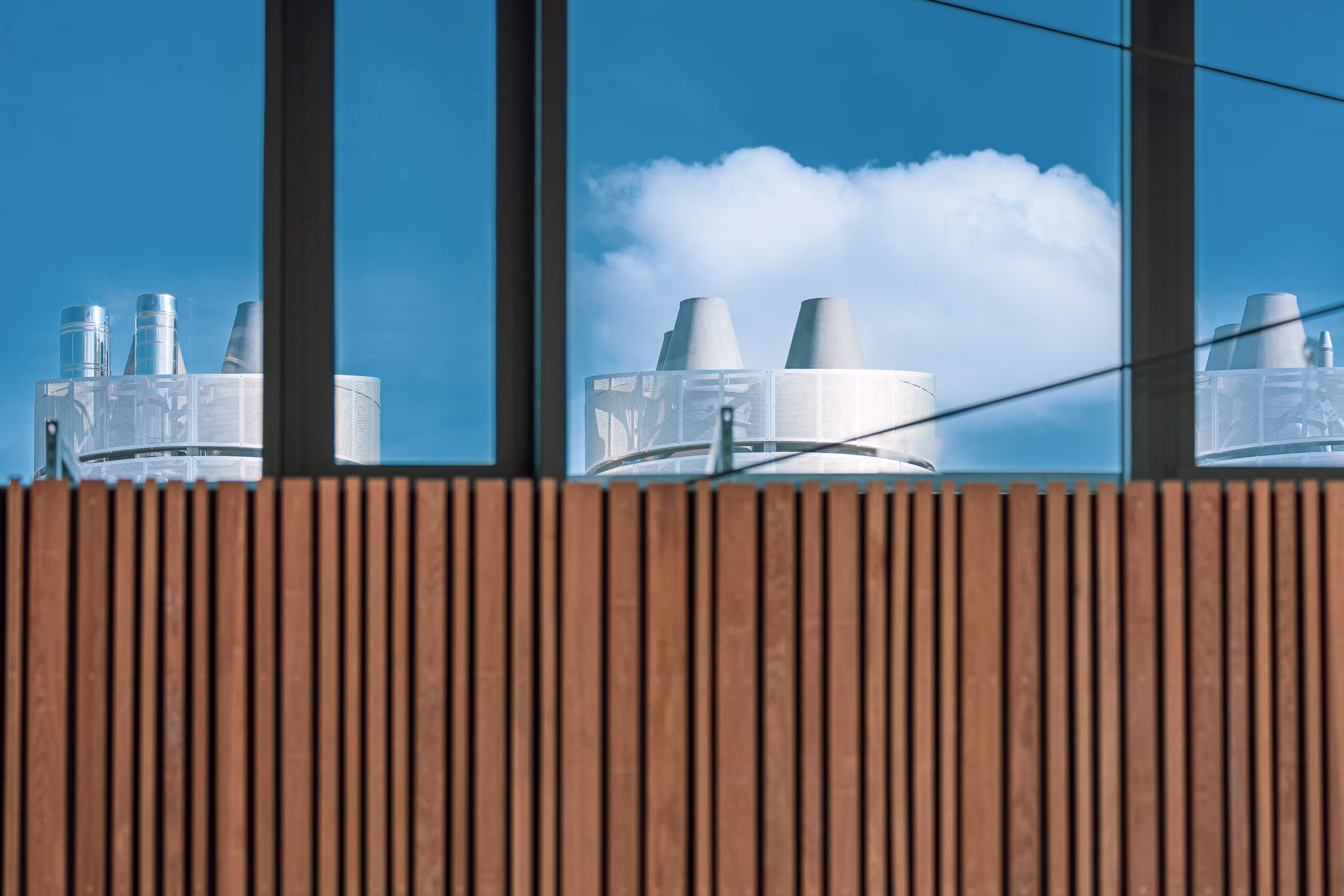 Reflections of industrial ventilation pipes on blue sky and clouds in a large window above vertical wooden slats.