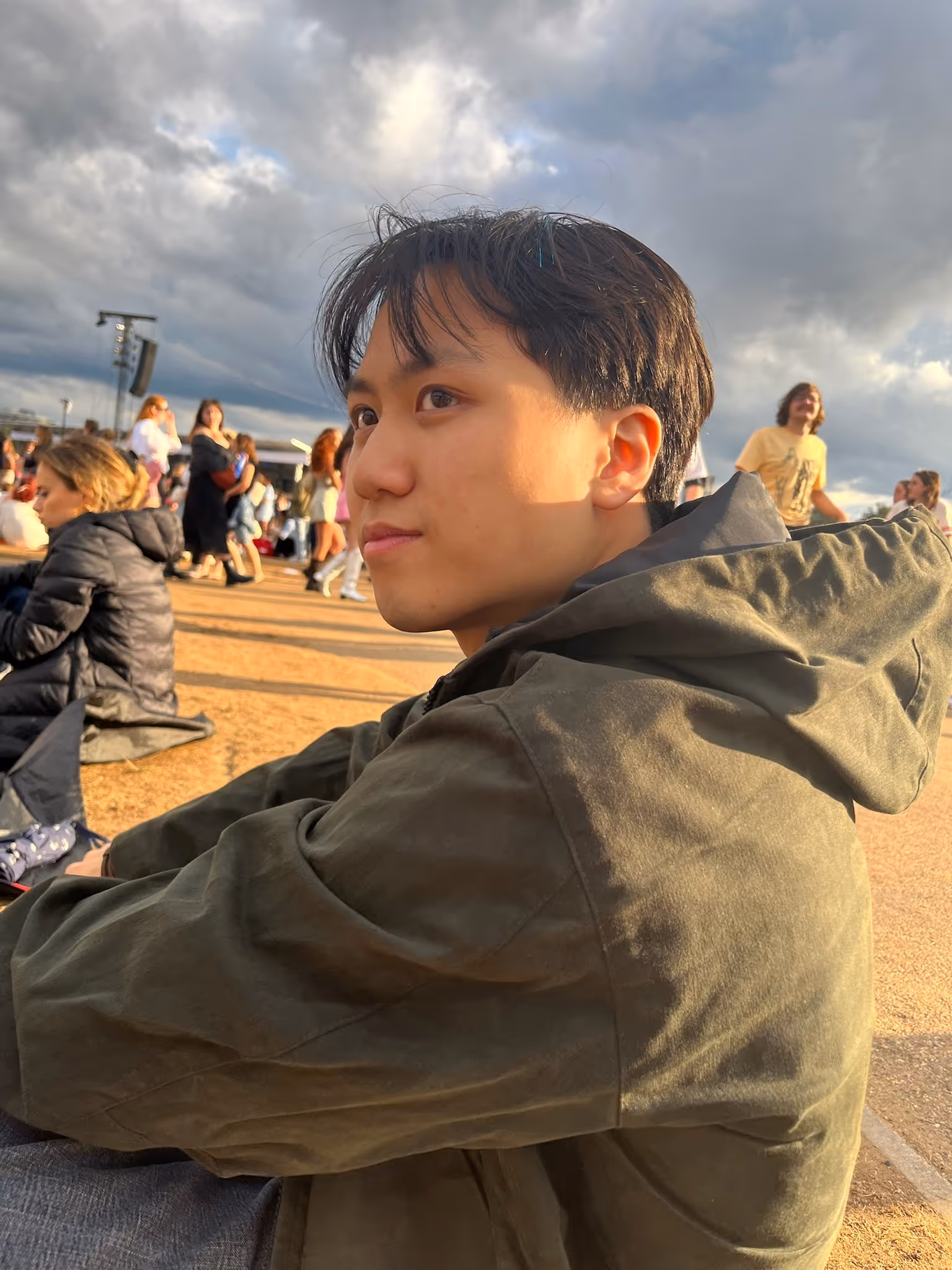 Young man in olive green jacket sitting outdoors on sandy ground during a cloudy day with people in the background.